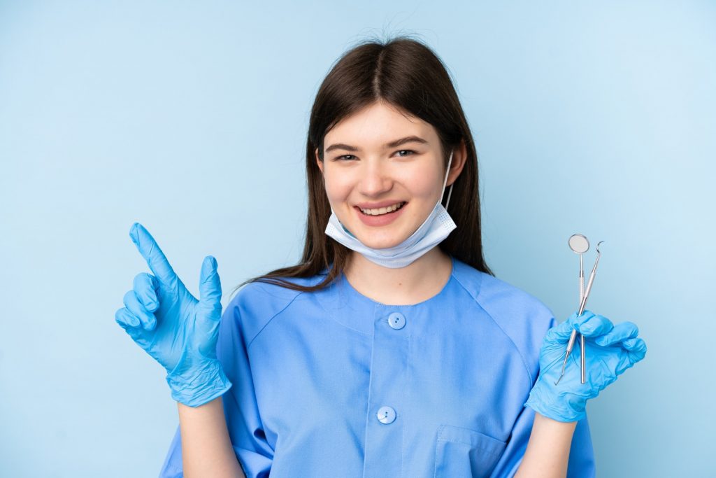 Young dentist woman holding tools over isolated blue background pointing finger to the side